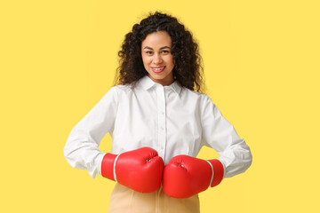 Young African-American businesswoman with boxing gloves on yellow background. Knockout concept