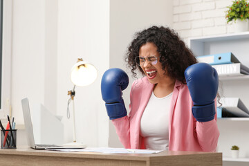 Angry African-American businesswoman with boxing gloves at table in office. Knockout concept