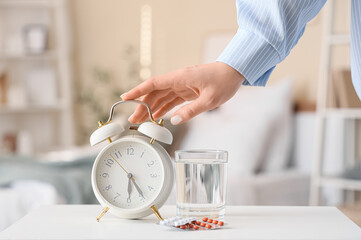Woman turning off alarm clock on bedside table with pills and glass of water in bedroom, closeup