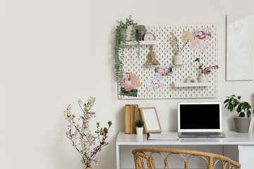 Workplace with blank laptop, blossoming branches and pegboard on light wall in office
