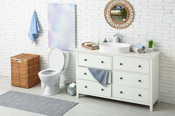 Interior of stylish restroom with ceramic toilet bowl, dresser and wicker basket