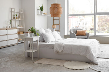 Interior of bedroom with cozy bed, grey armchair, houseplants and bedside tables