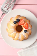 Tasty Bundt cake with powdered sugar, berries and server on pink wooden table, top view