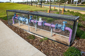 A recycled water irrigation system at a public park or sports field, featuring purple pipes, multiple valves, and a water meter enclosed in a wire mesh protective cage. Melbourne, Australia