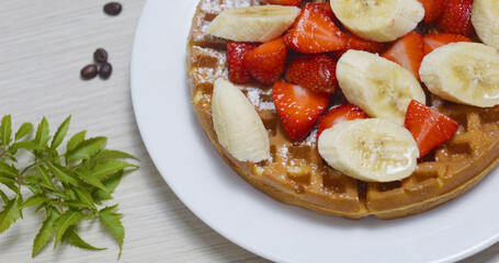 Crispy golden waffle topped with ripe strawberries, banana slices, powdered sugar sprinkle. Fresh mint leaves, coffee beans enhancing delectable breakfast presentation