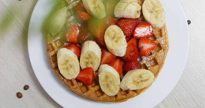 Golden waffle resting on pristine plate, topped with sliced bananas, fresh strawberries, dusted with powdered sugar, presenting delectable morning indulgence