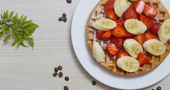 Crispy golden waffle garnished with sliced strawberries and bananas, sprinkled with powdered sugar, nestled on white ceramic plate near scattered coffee beans - Powered by Adobe