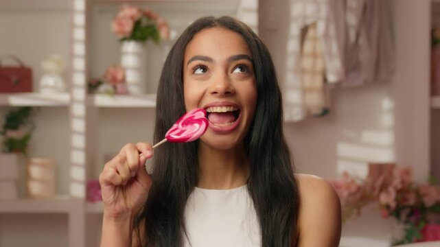 Beautiful latina woman girl holding candy lollipop funny smiling Hispanic teenager female in pink bathroom looking at camera eat bite sweets sugar toothy smile diet healthcare dental oral care beauty