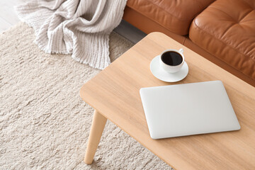 Modern laptop and cup of coffee on wooden coffee table in living room, closeup