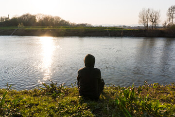 A lonely hooded, person, back to camera. Sitting by a river bank on a winters afternoon in the countryside