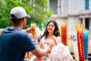 Young happy Latina in a traditional coral and white dress smiles brightly while receiving loaded fries with salsa at a street food stand in Alameda Central near Palacio de Bellas Artes, CDMX