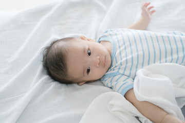 A three-month-old baby in a striped onesie lies peacefully on a soft white blanket, gazing up with wide, curious eyes, tiny hands waving, radiating innocence, comfort, and gentle charm.
