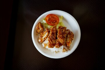 A plate of food with rice and meat on it. The meat is covered in sauce and the rice is white. There is a tomato on the plate and a leaf of lettuce