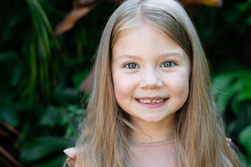 Fototapeta premium Smiling girl with long blonde hair and blue eyes standing outdoors in natural light. Happy childhood portrait with tooth gap. Real candid expression in nature setting, perfect for family themes.