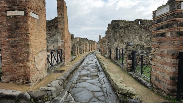 Pompeii, Italy - 8 January 2025. View of a paved Roman street, flanked by brick and stone ruins, with visible insula (block) numbers and iron railings marking the ancient building boundaries.