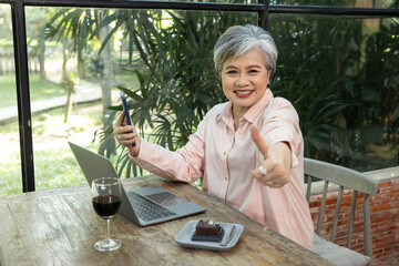 Cheerful elderly Asian woman with short gray hair wearing pink shirt holding smartphone and giving...