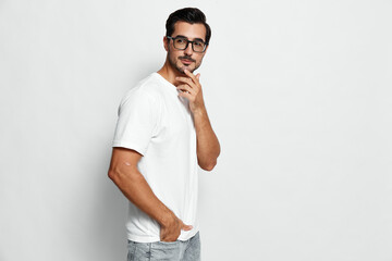 Fototapeta premium Confident young caucasian man in white t-shirt and glasses standing thoughtfully with hand on chin against a plain light background. Studio portrait concept.