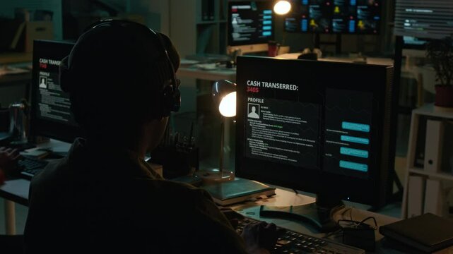 Rear over-shoulder shot of anonymous male scam call center operator wearing beanie and headset typing on computer in dark office while communicating with victim through online chat