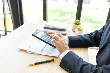 A man in a suit is sitting at a table with a piece of paper in front of him. He is looking at the paper and calculating and comparing data on the document, his phone, and a calculator.

