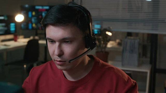 Close-up of face of focused young Asian male scam caller working at computer while speaking on internet phone to victim, impersonating government official to extort savings and money from bank account