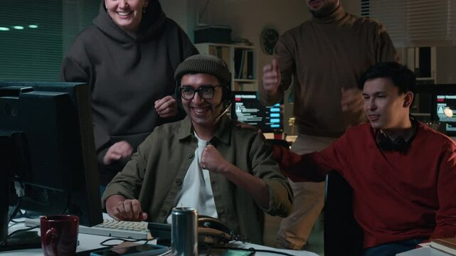 Medium shot of obnoxious multiethnic accomplices standing around Indian guy typing on computer, attentively looking at screen, then cheering at scamming victim of life savings in identity theft