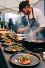 A chef preparing food in a pan with plates of appetizers lined up on a table for an event catering