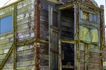 old wooden railway carriage in a severely deteriorated condition. The surface shows rotting boards, peeling paint, and rusted metal parts