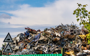 large pile of scrap metal composed of crushed and broken parts, including car and machinery fragments. In the background, blue sky and trees. The scene shows a metal scrapyard and recycling area