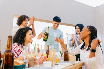 At table with food and drink on terrace, happy Latin friends celebrate with champagne