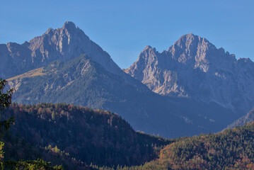 Majestic mountains rise above a vibrant autumn forest in Schwangau, Germany. Sunlight illuminates the rugged peaks, casting long shadows over the serene landscape.