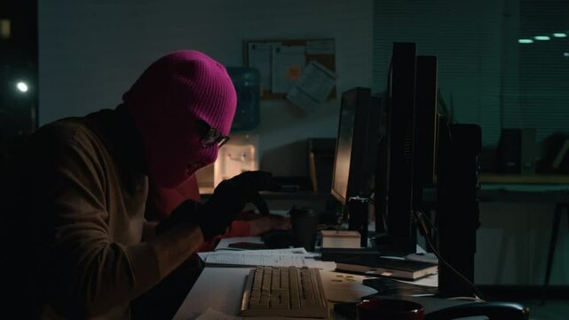 Medium shot of anonymous male scammer wearing pink balaclava and gloves typing on computer in office at night while launching online attack at victim, rubbing hands, turning to camera and gloating