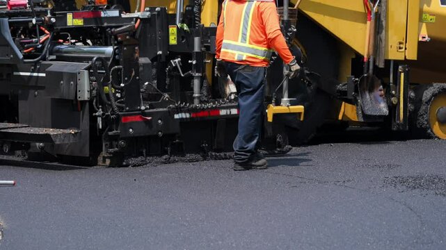 Asphalt paving machine laying fresh blacktop on a road. A worker in a high-visibility vest is taking a shovel.