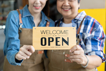Asian senior woman and young woman wearing aprons smiling while holding wooden open welcome sign together in front of coffee shop symbolizing teamwork partnership and hospitality in family business