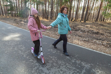 Little girl riding scooter while walking with her grandmother in the forest on a sunny day
