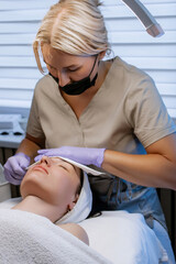 Esthetician cleansing woman's face with towel after facial treatment in beauty salon, young woman relaxing with closed eyes