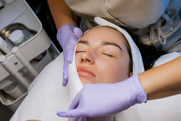 Esthetician cleansing woman's face with towel after facial treatment in beauty salon, young woman relaxing with closed eyes