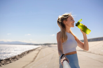 Smiling young blonde woman jogger runner drink water from bottle at river embankment closeup