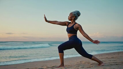 A mature black woman practices yoga on a sandy beach during sunrise. She is in a warrior pose, wearing sportswear. The ocean waves gently lap at the shore. Active lifestyle - Powered by Adobe