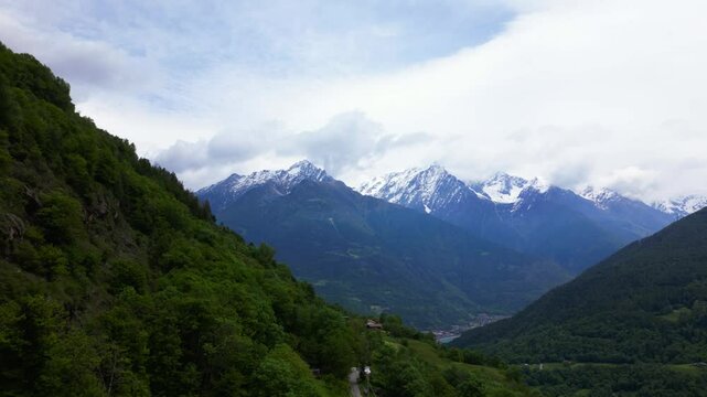 Sweeping drone view of snow-covered peaks and lush green valleys near Edolo, with dramatic clouds and layered mountain ridges. Aerial view from Vico, Edolo, Italy Italia