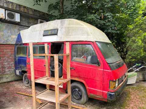 A worn-out abandoned red caravan van, covered in moss and dirt, is parked next to a green and stone building facade.
