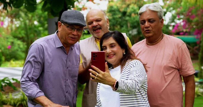 Senior friends video calling together on smartphone, cheerful Indian Asian elderly group standing outdoors in lush green garden, waving, smiling during mobile video communication with loved ones