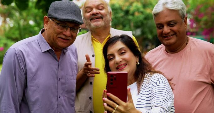 Senior friends video calling together on smartphone, cheerful Indian Asian elderly group standing outdoors in lush green garden, waving, smiling during mobile video communication with loved ones