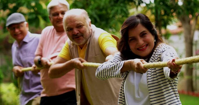 Senior playing tug of war in garden, Indian Asian elderly friends enjoying outdoor physical activity with rope, laughing and pulling together as part of fun reunion game amid green nature