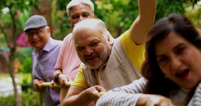 Senior playing tug of war in garden, Indian Asian elderly friends enjoying outdoor physical activity with rope, laughing and pulling together as part of fun reunion game amid green nature