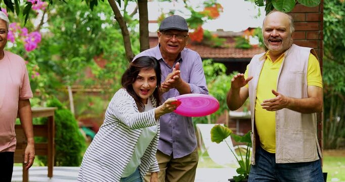 Senior friends enjoying throwing frisbee outdoors, Indian Asian elderly group playing with flying disc in garden, smiling and laughing together while celebrating reunion in lush green nature setting