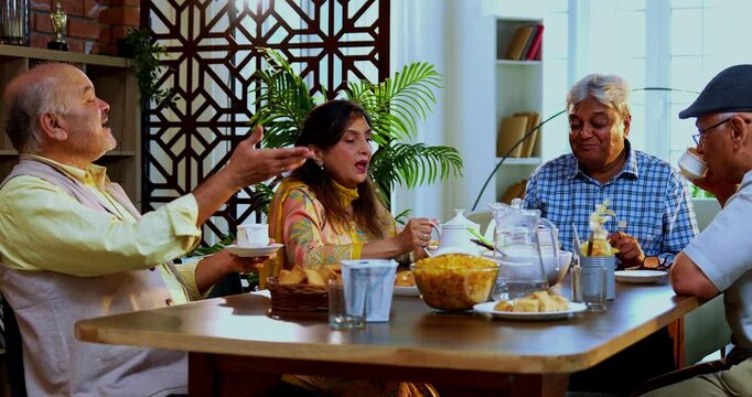 Seniors serving and eating together, Indian Asian old age friends reuniting at home eating food, serving snacks, cold drink, tea or coffee to each other while seated at modern dining table, enjoying