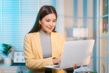 Asian woman works overtime at her office desk late at night, surrounded by paperwork, laptop, and smartphone, showing resilience and focus as she manages business tasks in a quiet