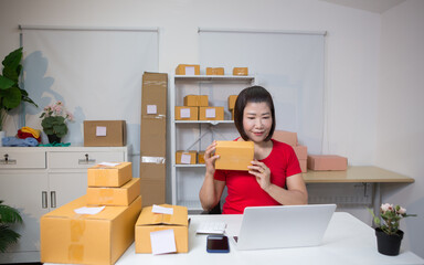 An Asian woman in a light red T-shirt is selling goods online, efficiently packing orders for...