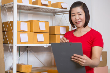 An Asian woman in a light red T-shirt is selling goods online, efficiently packing orders for...