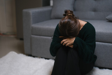 A young woman, who appears to be of Asian descent, sits curled on the floor with a melancholic...